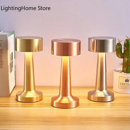 Three metallic Costa Rica Coffee Friends LED Table Lamps Retro Bar Coffee Table Lamp Rechargeable Touch Sensor Wireless Night Lights with cylindrical bases and wide, flat tops illuminate a wooden surface next to a cactus plant and several books, including one titled &quot;Les Miserables.&quot; The lamps, featuring LED touch sensors, are gold, copper, and silver in color.