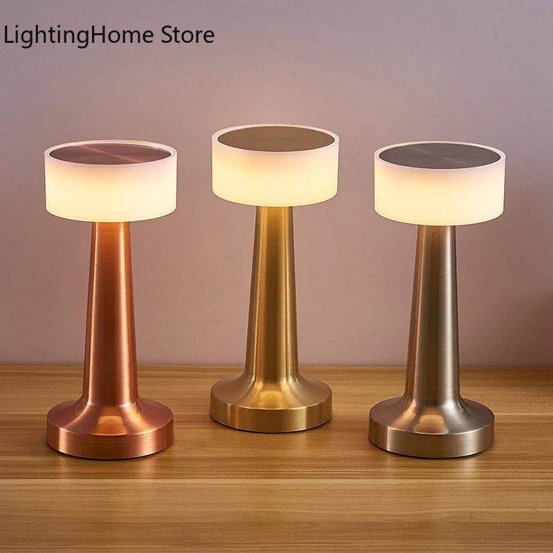 Three LED Table Lamps from Costa Rica Coffee Friends, featuring a sleek metallic design and cylindrical bases, sit on a wooden surface. The Retro Bar Coffee Table Lamps, in copper, gold, and silver finishes, have round diffusers at the top, casting a warm, ambient light. The backdrop is a plain, light-colored wall.