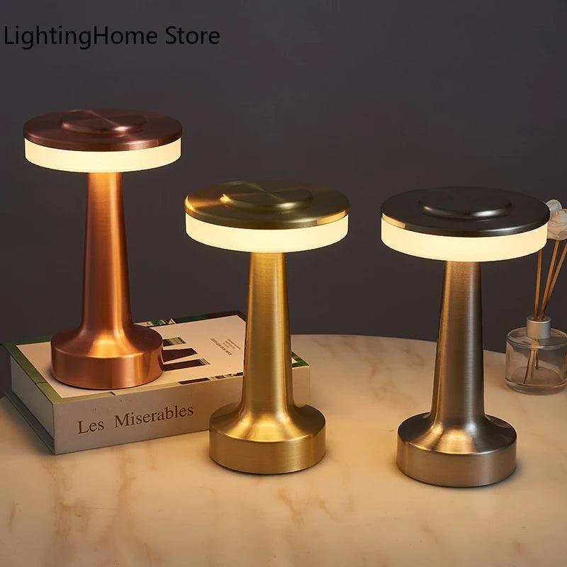 Three LED table lamps from Costa Rica Coffee Friends, featuring cylindrical bases and round lighted tops, are displayed on a table. These rechargeable touch sensor wireless night lights come in copper, brass, and black finishes. The table also holds a stack of books, including a visible copy of &quot;Les Miserables,&quot; and a small decorative piece.