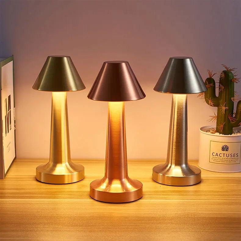 Three retro table lamps from Costa Rica Coffee Friends, available in metallic finishes of gold, copper, and silver, are elegantly displayed on a wooden surface. Each &quot;LED Table Lamp Retro Bar Coffee Table Lamp Rechargeable Touch Sensor Wireless Night Light Restaurant Living Room Decor Light&quot; features a conical shade and emits a warm glow. A cactus plant in a white pot on the right side adds a refreshing touch of greenery.