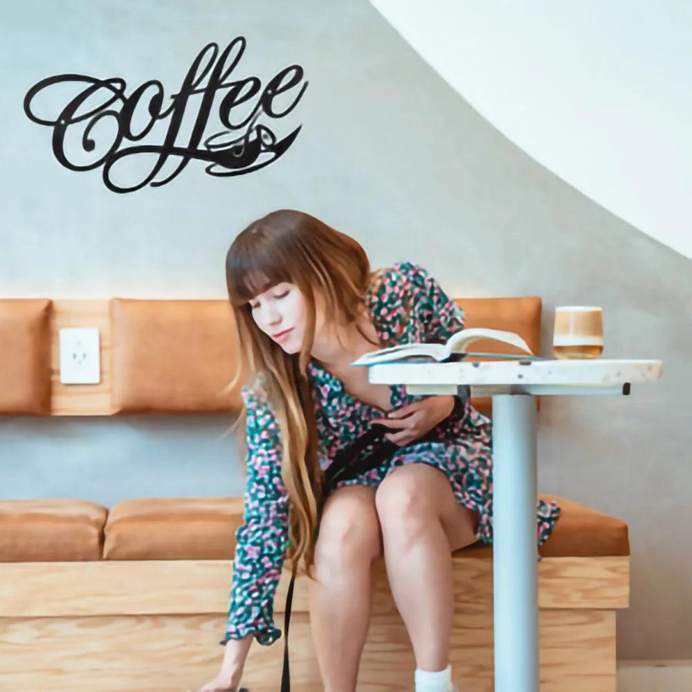 A woman with long hair and a floral dress sits at a modern café with wooden furniture. She reaches down with her left hand while sitting on a bench seat, an open book and a glass of coffee in front of her on the table. The word &quot;Coffee,&quot; artistically portrayed as the COFFEE Metal Word Cafe Wall Sign Hanging Kitchen Black Plaque Art Home Decoration Bar Plaque Silhouette Decor by Costa Rica Coffee Friends, decorates the wall.