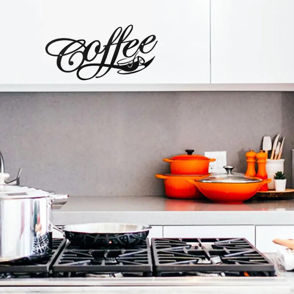 A kitchen features a stove top and pots. Above the counter is a white cabinet adorned with the Costa Rica Coffee Friends COFFEE Metal Word Cafe Wall Sign Hanging Kitchen Black Plaque, which artistically displays the word &quot;Coffee&quot; in black cursive. On the counter are orange pots, a small plant, and some cooking utensils. A piece of metal wall art enhances the home decoration above the grey backsplash.