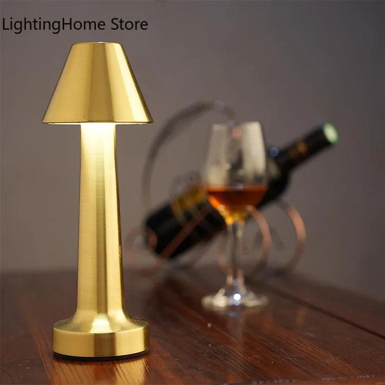 A sleek, gold LED Table Lamp Retro Bar Coffee Table Lamp Rechargeable Touch Sensor Wireless Night Light by Costa Rica Coffee Friends illuminates a wooden surface. In the background, there is a glass of amber liquid and a bottle of wine lying on its side in a wire wine holder. The lamp casts a soft, warm light, creating a cozy ambiance. The text &quot;LightingHome Store&quot; is in the top left corner.